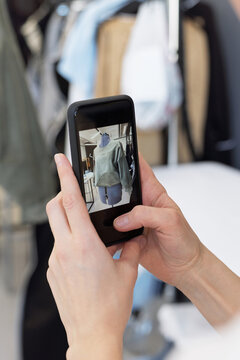 Close-up view of unrecognizable woman holding smartphone and taking picture of stylish garment on mannequin in tailor shop