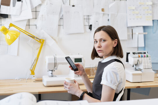 Portrait of young female tailor looking at camera while resting in atelier with coffee and smartphone on background of garment sketches