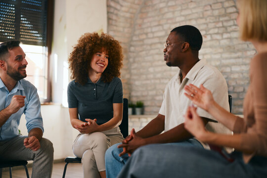 Smiling young woman with curly hair looks at an African American man speaking expressively during a positive and supportive multi-ethnic group therapy session indoors.