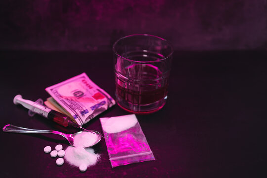Neon-lit scene with syringe, white pills, powder, money banknotes and alcohol drink glass on dark background with copy space, concept of drug addiction, substance abuse and nightlife risks