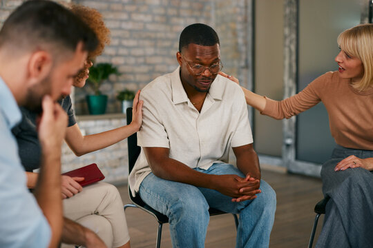 Vulnerable African American man receiving comforting support and touch from two women during an intimate multi-ethnic group therapy or counseling session indoors.