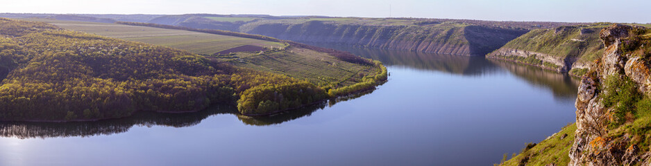 A breathtaking panoramic view of the Dniester River canyon near Subich, Ukraine. Mossy rocks are in the foreground, overlooking the calm blue water and green forested hills under a bright sun.