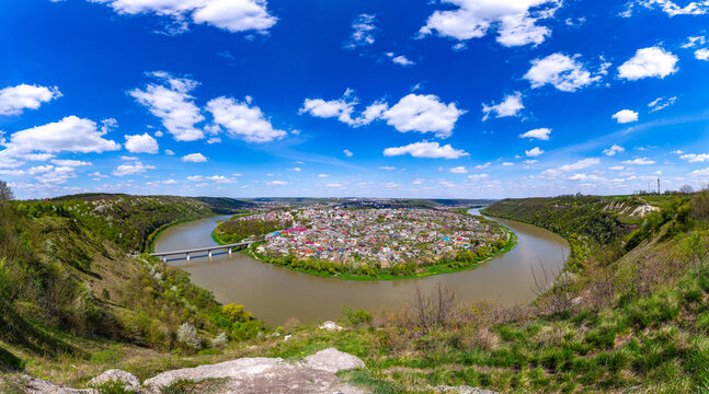 A stunning high-angle panoramic view of Zalishchyky, Ukraine, located in a dramatic meander of the Dniester River. The town is surrounded by steep green hills under a bright blue sky with white clouds - Powered by Adobe