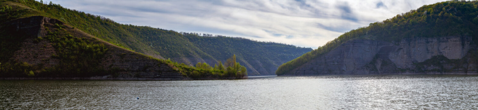 A beautiful panoramic landscape of the Ternava River canyon, a tributary of the Dniester in Ukraine. Steep, forested hills and rocky cliffs surround the calm, rippling water.