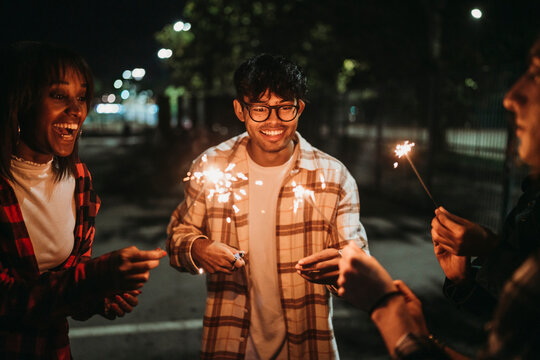 Positive young multiracial friends in casual clothes smiling and looking at burning sparklers while standing on city street at night