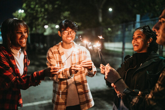 Positive young multiracial friends in casual clothes smiling and looking at burning sparklers while standing on city street at night