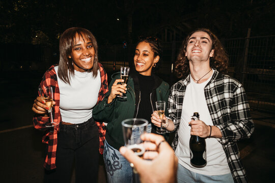 Group of happy young people in casual clothes smiling and looking at camera with glasses of champagne at night