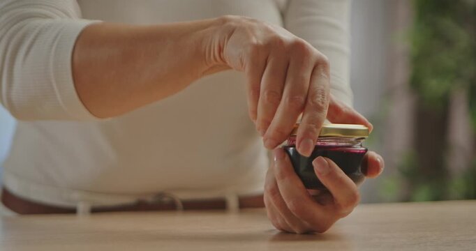 Close up of female hands twisting open a small glass jar with golden lid containing dark jam or sauce, on wooden table in soft natural light. Slow motion.