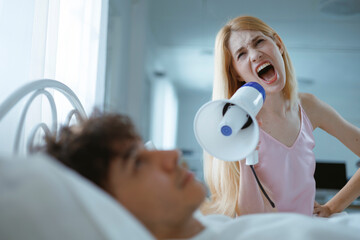 A white woman screams into a megaphone at her sleeping husband, trying to wake him up in the...