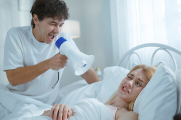A white man shouts into a megaphone at his sleeping wife, trying to wake him up in the morning.