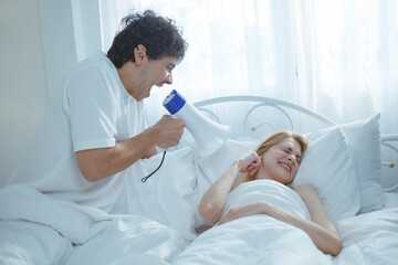 A white man shouts into a megaphone at his sleeping wife, trying to wake him up in the morning.