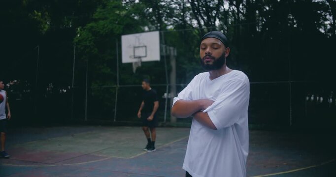 Basketball player of African descent in oversized white shirt and durag standing with arms crossed on outdoor court, confident athletic stance