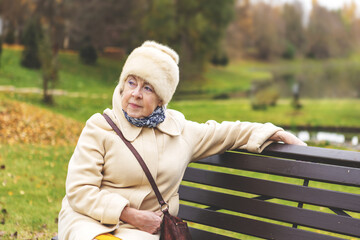 Fototapeta premium Thoughtful senior woman sitting on park bench surrounded by autumn nature. Elderly female enjoying outdoor, peaceful aging and natural beauty in fall season