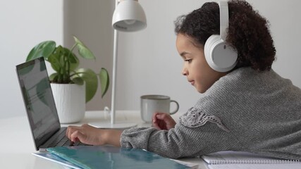 Primary school African American Student child with headphones hand writing in book using laptop. Distance learning online education. School girl studying at home with digital tablet computer and doing - Powered by Adobe