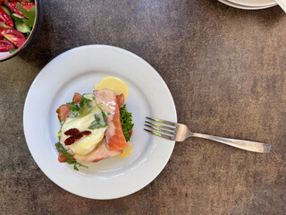 An elegant brunch plate featuring eggs Benedict with smoked salmon, hollandaise sauce, and fresh greens on a white plate next to a silver fork on a dark brown table.