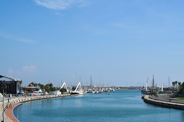 the harbor of Valencia, Spain. panoramic view. blue water and sailboats in a distance. clear blue sky. green palm trees. sidewalk for pedestrians. industrial building. summer scene. travel and tourism