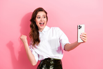 Young woman celebrating success while taking a selfie with a smartphone against a pink background...