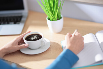 Woman with notebook and pencil having coffee break at wooden table indoors, closeup