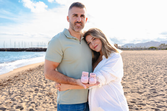 Expecting couple standing on sandy beach, smiling at camera, pregnant woman holding tiny pink baby shoes with love and tenderness. Bright sky, calm sea and sunlight create peaceful,emotional maternity - Powered by Adobe