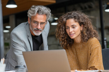 Two diverse business professionals, a mature man and a young woman, collaborate intensely, reviewing content on a laptop in a modern office. They are engaged in a productive discussion.