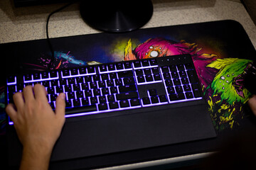 Close up of gaming keyboard with RGB backlighting and colorful gaming mousepad, showing hand of gamer, highlighting modern gaming peripherals in intense gaming setup