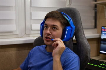 Close up of young man wearing blue gaming headphones and using microphone while sitting at gaming desk. Scene highlights gaming environment, featuring person in relaxed, focused posture