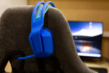 Blue gaming headphones hangs on back of gaming chair. The colorful design contrasts with the subtle background of computer screen, evoking sense of preparation for intense gaming action
