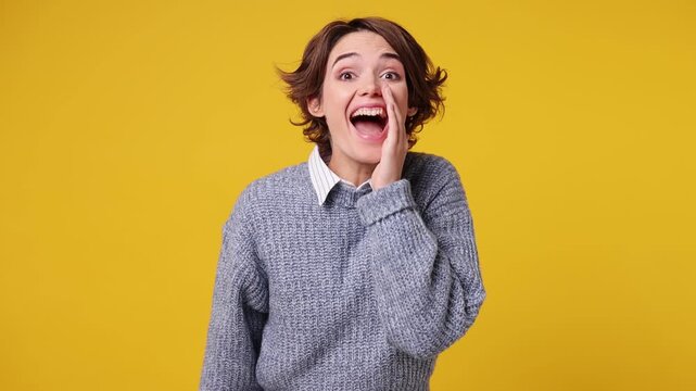 Excited happy young woman wear grey knitted sweater white shirt casual clothes scream hot news about sales discount with hand near mouth hurry up isolated on plain yellow background. Lifestyle concept
