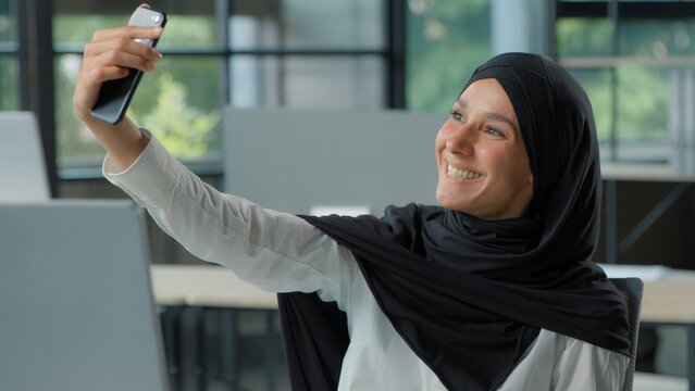 Smiling happy Arabian Muslim business woman in hijab take selfie photo with phone in office Islamic businesswoman female girl posing at workplace photographing with smartphone mobile social media vlog