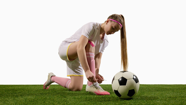 Focused female football player kneeling on grass tying shoes with ball beside. Concept of preparation, concentration, responsibility, dedication in sports lifestyle and education. - Powered by Adobe
