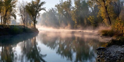 Fototapeta premium River fog forming over water surface at sunrise, reflecting green and golden trees along riverbanks in autumn