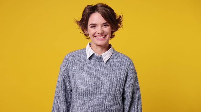 Smiling friendly charming young woman she wearing grey knitted sweater white shirt casual clothes looking camera wink eye blink isolated on plain yellow background studio portrait. Lifestyle concept