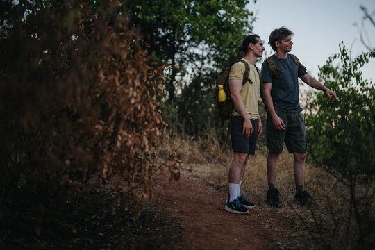Two friends hike along a dry, rugged trail at dusk, planning their next move. They wear casual outdoor gear and backpacks, surrounded by trees and warm natural tones.