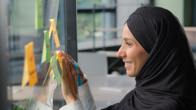 Smiling Muslim business woman stick sticky notes on glass in modern office Arabian businesswoman in hijab preparing planning work tasks female girl corporate leader leaving note organize job strategy