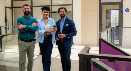 Three smiling business people standing together in a modern office building corridor, enjoying a coffee break and representing teamwork and success