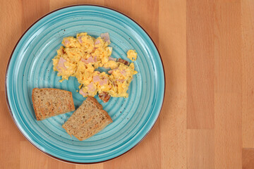 Overhead shot of a breakfast plate with creamy scrambled eggs mixed with ham and two slices of whole grain toast, set on a light wooden table
