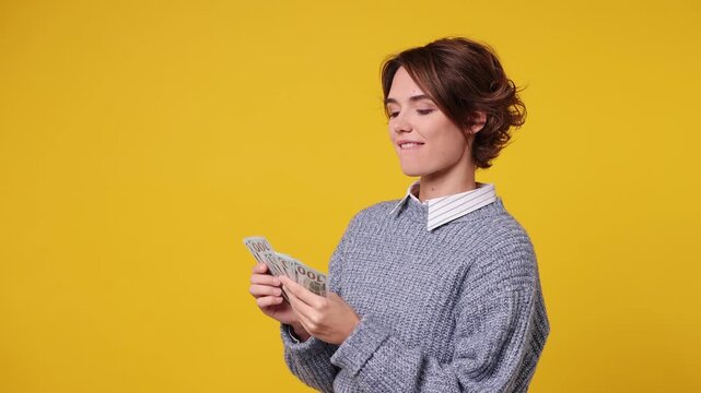 Happy accountant young woman she wear grey knitted sweater white shirt casual clothes hold in hand count stack of cash money in dollar banknotes isolated on plain yellow background. Lifestyle concept