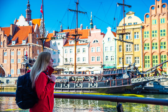 Tourist in red jacket with backpack walks along Gdansk waterfront. Behind her are historical buildings with vibrant facades and a traditional sailing ship at the harbor.