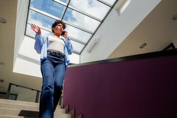 Young businesswoman gesturing while making a phone call and climbing the stairs in a modern office building, seen from a low angle perspective
