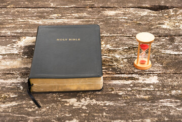 A top-down view (flat lay) of a closed black Holy Bible next to a vintage hourglass on a rustic wooden table in soft, natural morning light, symbolizing faith, time, and contemplation.