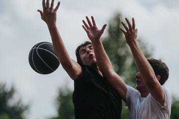 Two young men reach for a basketball in a tense outdoor game, highlighting competition,...