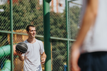 A young man stands on an outdoor basketball court, holding a ball and smiling. A chain-link fence and greenery frame the scene, suggesting a casual, sunny practice or pickup game. © qunica.com