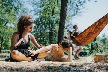 Three friends relax outdoors on a sunny day, one applying sunscreen while another chats, a person...