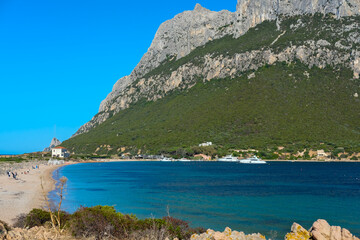 Scenic coastal view with clear turquoise water, a sandy beach, and a green mountain slope under a bright blue sky.