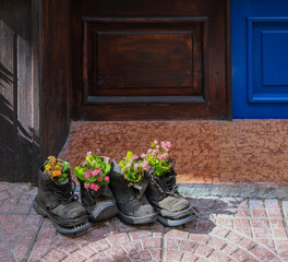 Old black boots repurposed as creative flower pots with blooming pink and orange flowers in front of a wooden door.