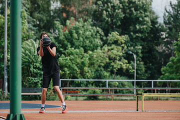 A focused young man in black athletic gear practices on an outdoor court, catching and shooting a basketball amid green trees, benches, and a hoop, conveying fitness and outdoor training.