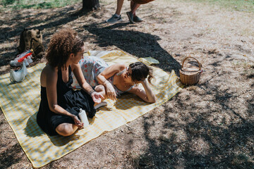 Two women share a relaxed moment on a sunny day, chatting on a yellow plaid blanket. They have a basket, water bottle, and snacks nearby in a leafy park setting.