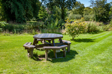 Several modern, round, grey outdoor tables with attached stools are set up on a lush green lawn, surrounded by trees in a park-like setting.