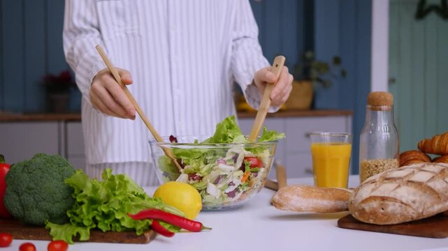 Woman in pajamas adding fresh lettuce into a glass bowl and mixing a healthy vegetarian salad with wooden spoons on a kitchen table with vegetables, bread, and juice in the background