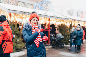 Young woman on christmas market. cute lovely girl in red hat standing at street and hangs ball on...